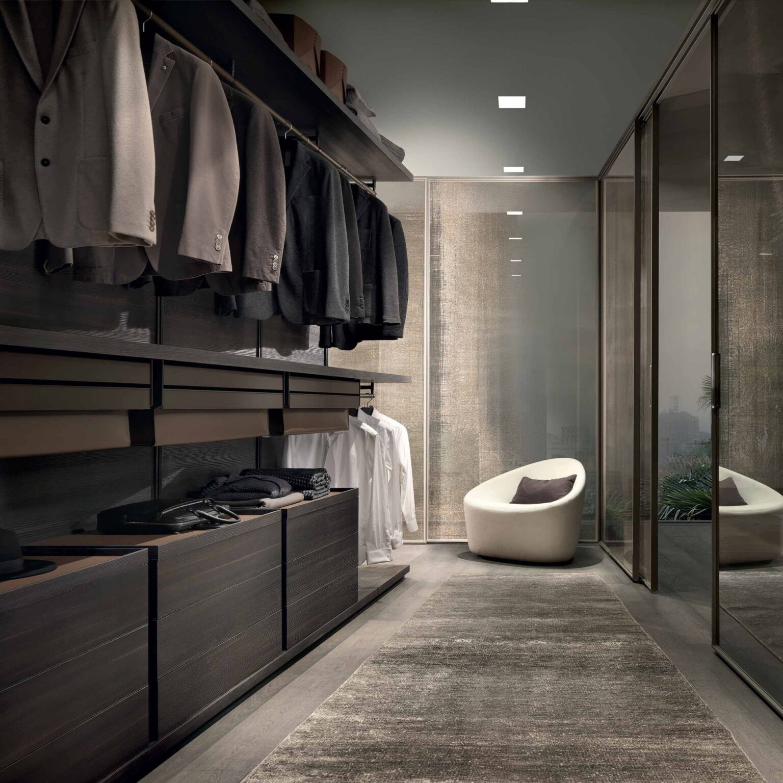 Inloopkast met open garderobesysteem langs wand en glazen scheiding met zicht op zitgedeelte in luxe dressingruimte
