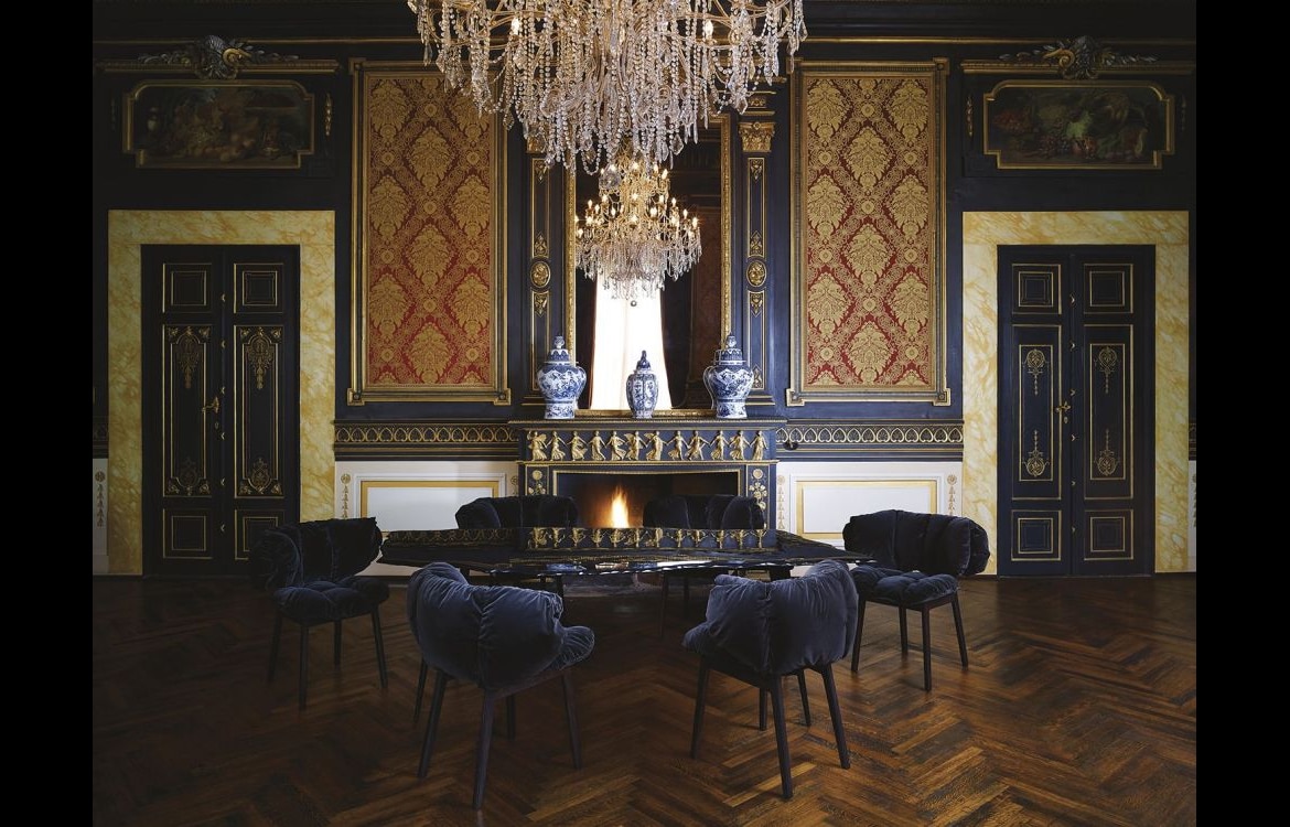 De Blue Velvet stoel in een klassiek interieur met een glazen tafel en antieke decoratie. De zwarte fluwelen bekleding geeft een verfijnde en luxueuze uitstraling aan het geheel, passend bij verschillende stijlen.