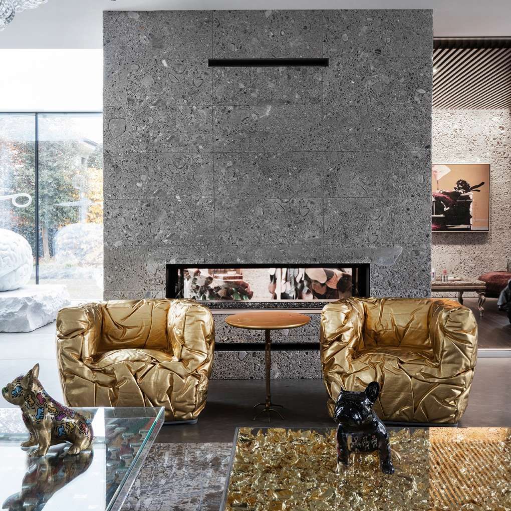 De Sponge-armchairs, uitgevoerd in een opvallende goudkleur, staan centraal in een eigentijdse en luxe ruimte met een natuurstenen muur en een moderne open haard. De combinatie van het goud en de grijze stenen achtergrond zorgt voor een contrastrijke, maar harmonieuze uitstraling. De sculpturale vormen en de onregelmatige rimpels in de leren bekleding van de stoelen benadrukken de unieke esthetiek en het vakmanschap van het ontwerp.