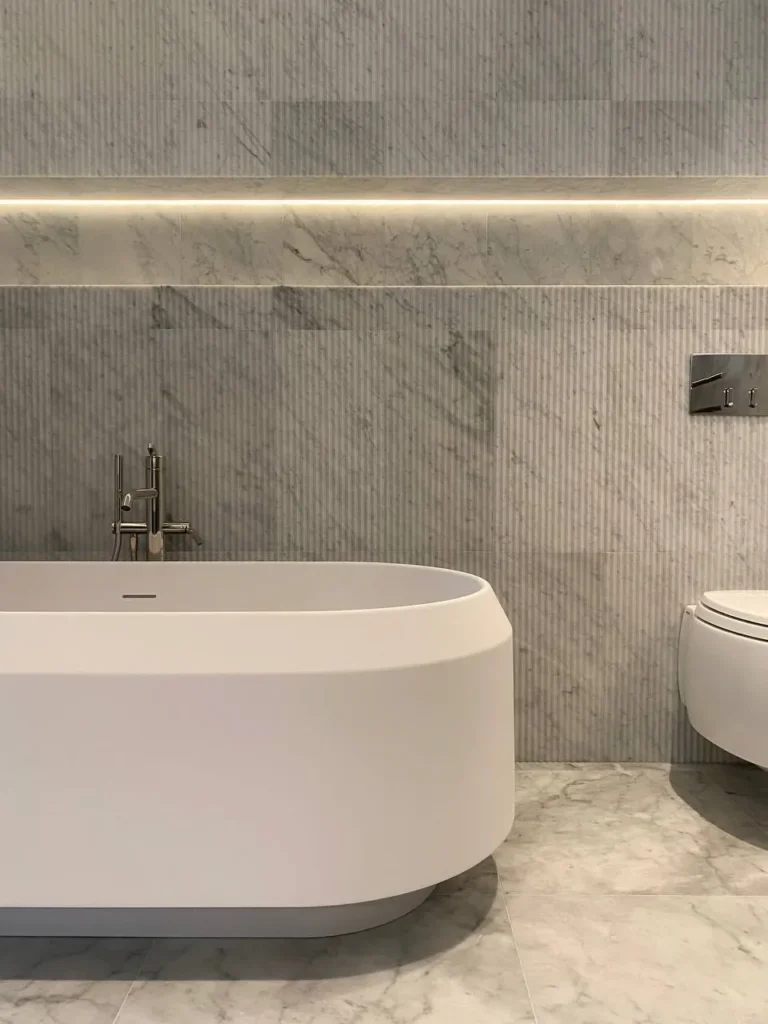 Agape vrijstaand bad in matte afwerking gecombineerd met Salvatori natuurstenen wandtegels met verticale ribstructuur en geïntegreerde wandverlichting in minimalistische badkamer